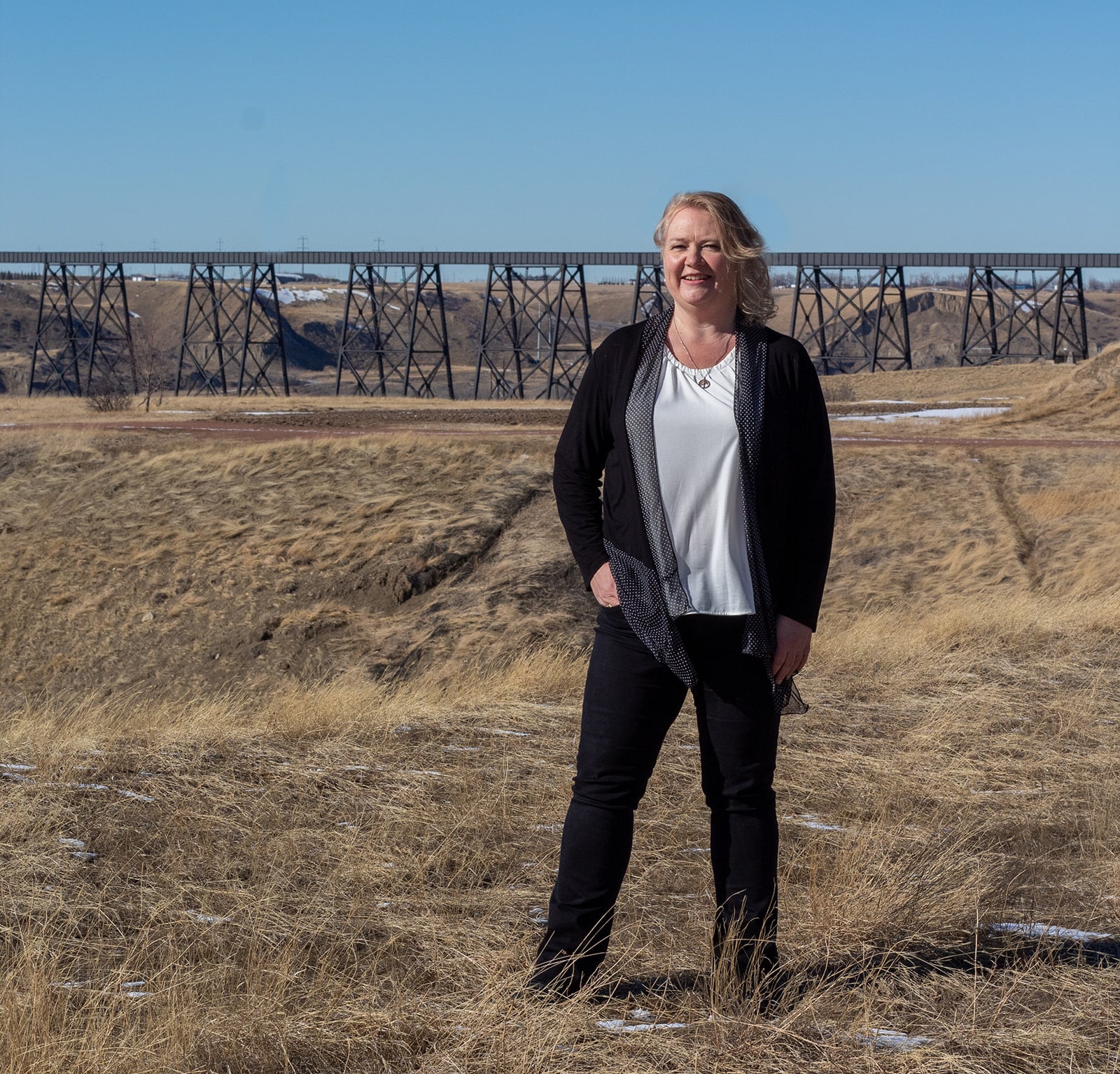 Smiling woman in black cardigan and white blouse stands in a dry grassy field with a long steel trestle bridge behind her under a clear blue sky.